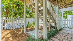 Una estructura de pérgola de madera con un columpio, rodeada por un cerco de piquetes blancos y abundante vegetación.
