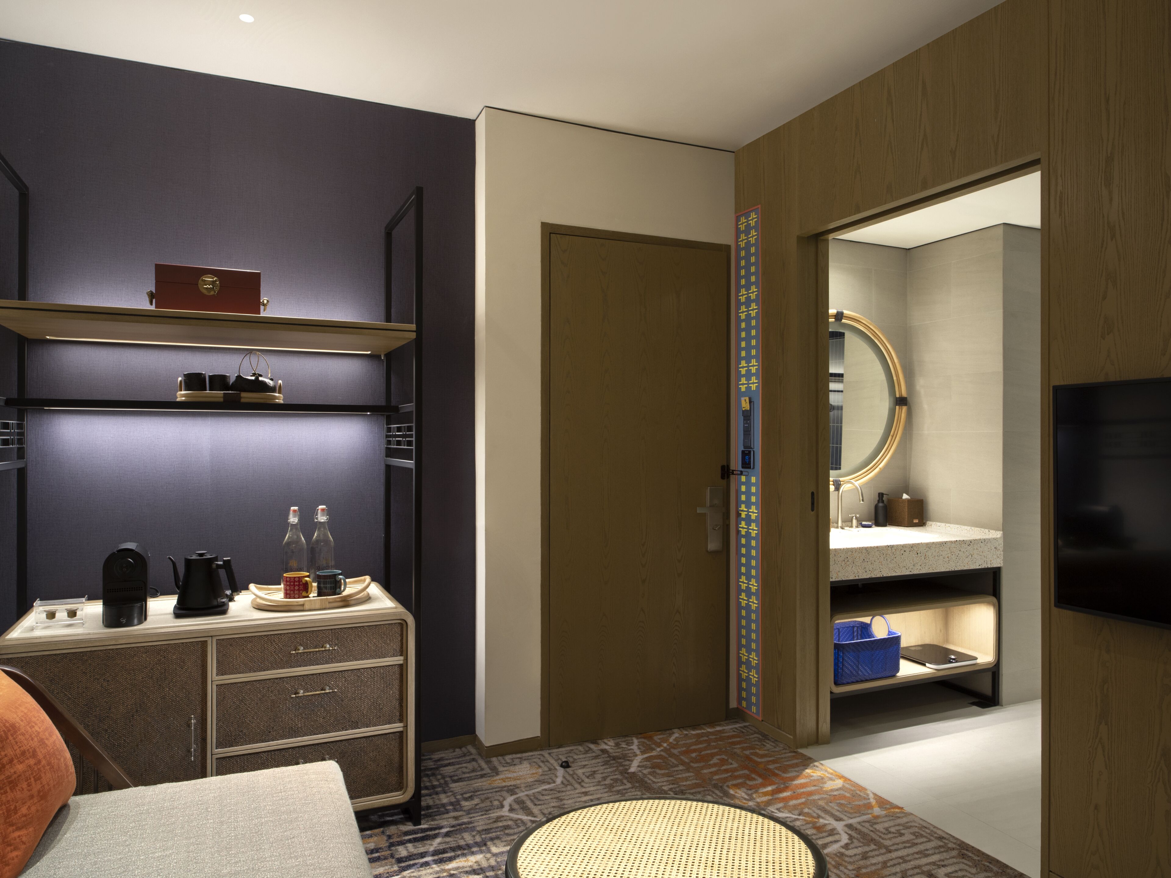 A modern hotel room with a dark blue accent wall, a wooden door, a small kitchenette, a television, and a bathroom with a mirror and sink.