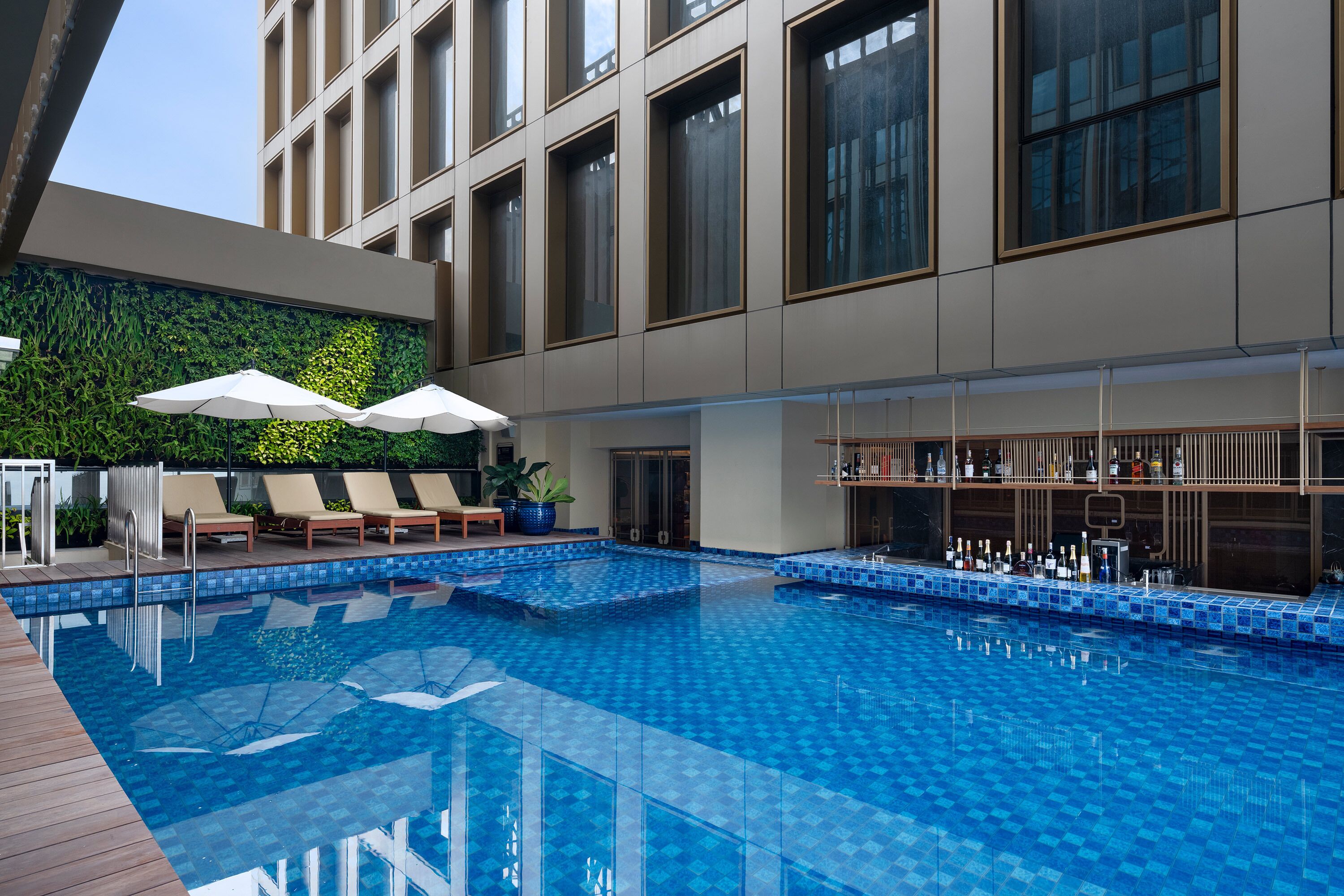 A modern indoor pool area with lounge chairs and umbrellas, a bar with a variety of bottles, and a green wall with plants.