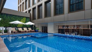 A modern indoor pool area with lounge chairs and umbrellas, a bar with a variety of bottles, and a green wall with plants.