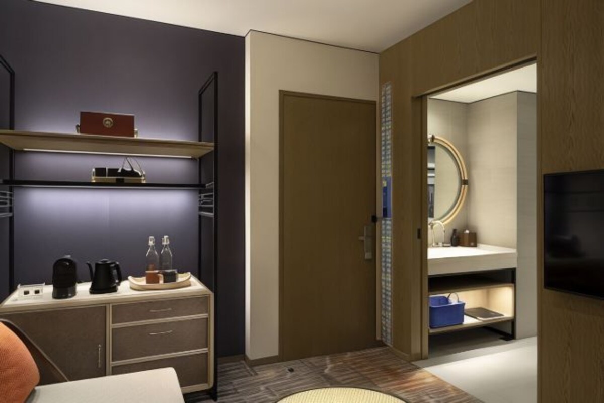 A modern hotel room with a dark blue accent wall, a wooden door, a built-in TV, a small kitchenette, and a large mirror.