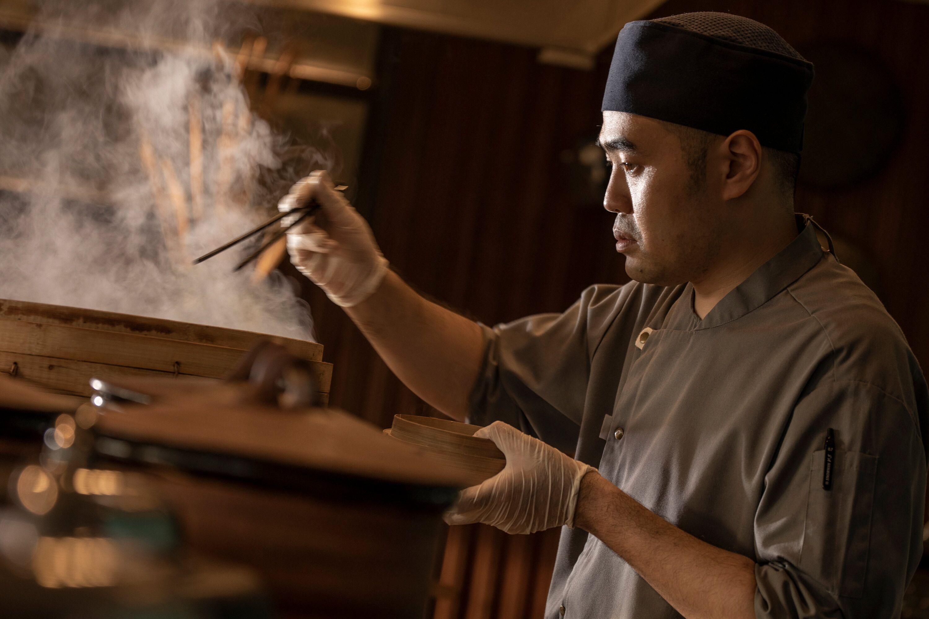 Una persona con uniforme sosteniendo palillos y una bandeja de madera, presumiblemente en una cocina.
