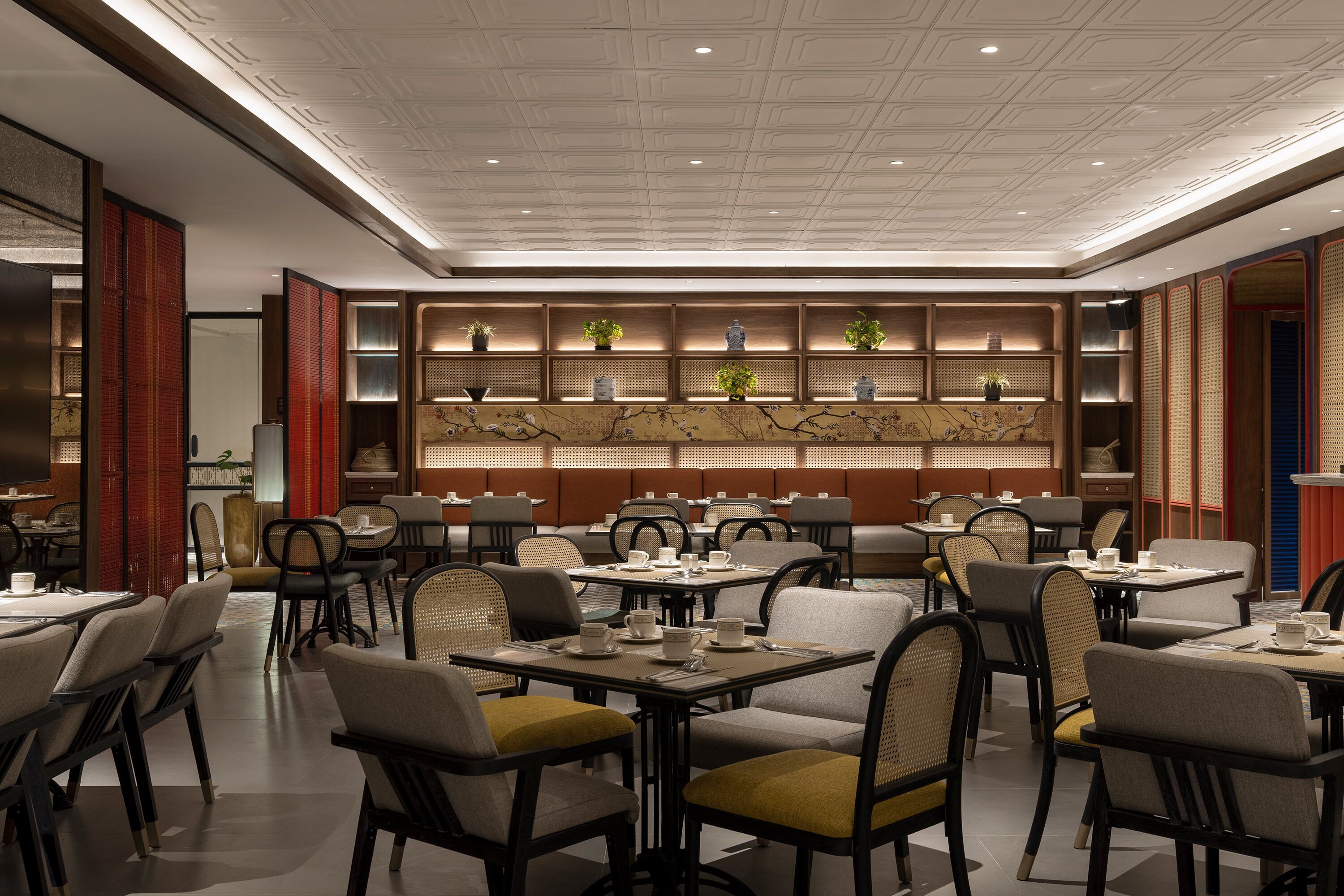 A modern restaurant with multiple dining tables and chairs, a decorative wall with shelves, and a ceiling with recessed lighting.