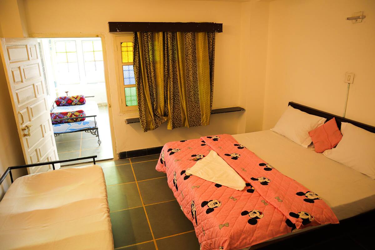 A room with two beds, one with a pink Mickey Mouse quilt and another with a white mattress. There is a window with curtains, a door leading outside, and a small table with a colorful bag.