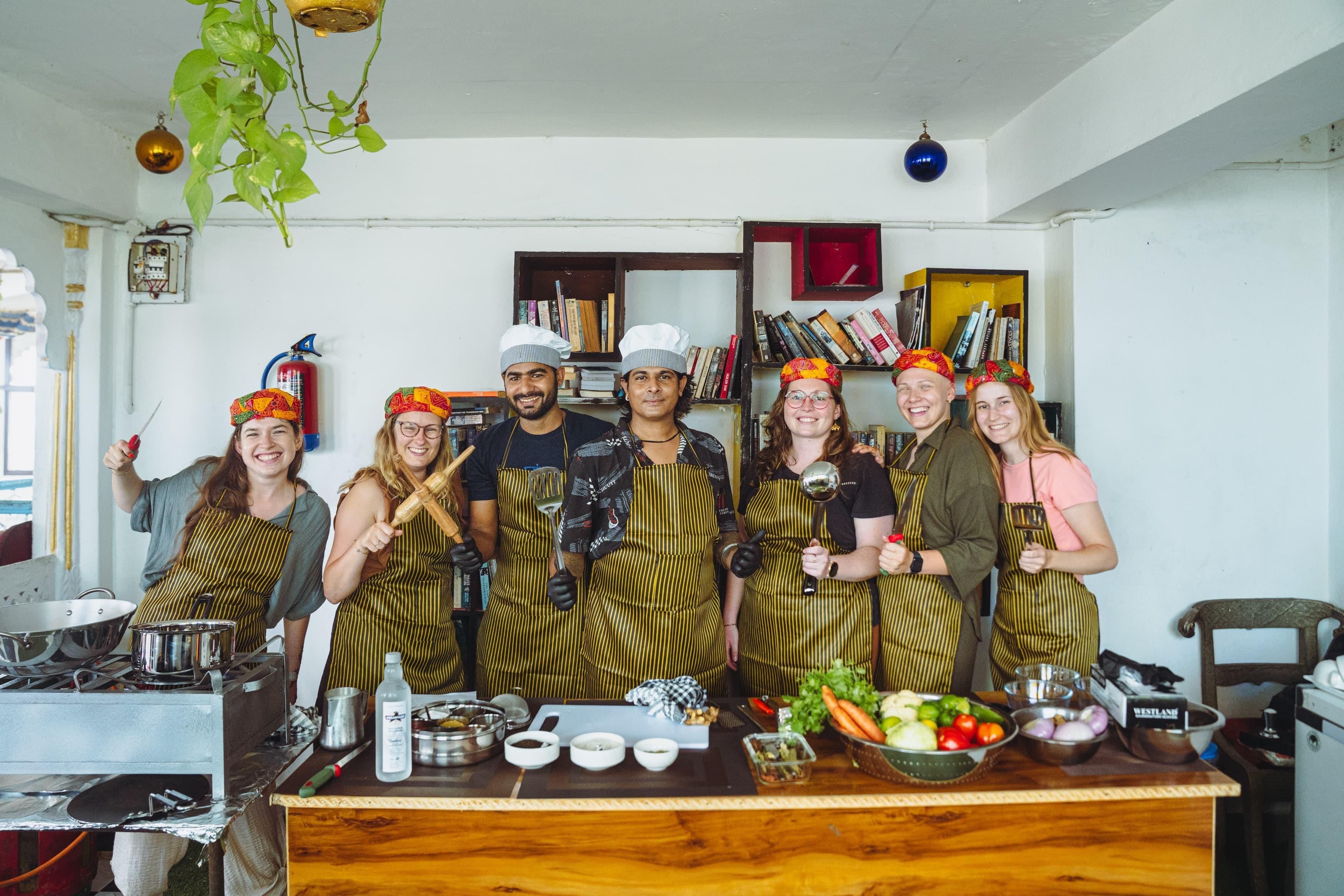 A group of people in a kitchen with striped aprons, a wooden table with cooking utensils, and a bookshelf in the background.