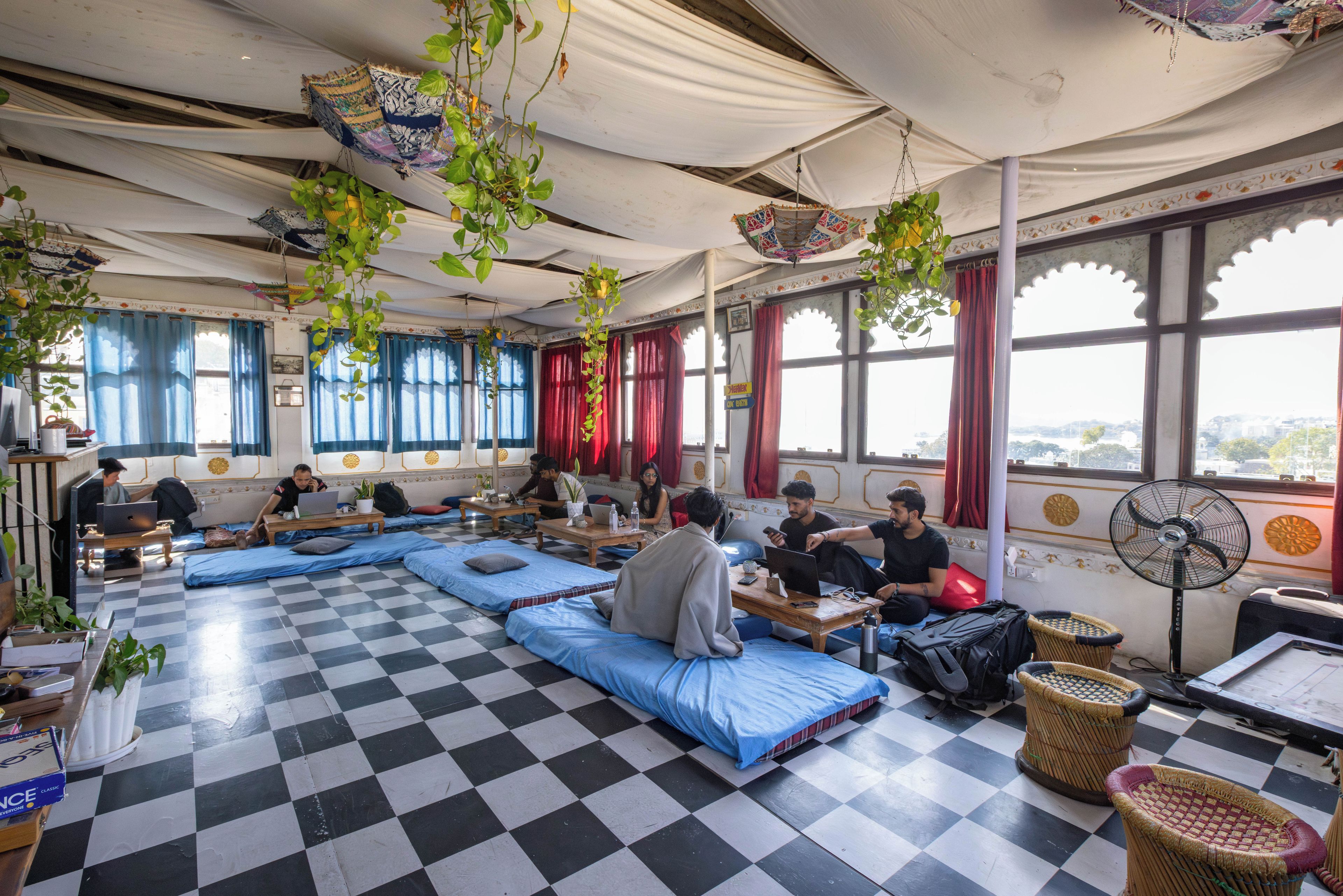Una habitación con suelo de cuadros, asientos acolchados azules, plantas colgantes y ventanas grandes con cortinas rojas.