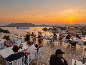 A terrace with tables and chairs overlooking a body of water and a sunset. - Dreamyard Udaipur - Hostel (Udaipur)