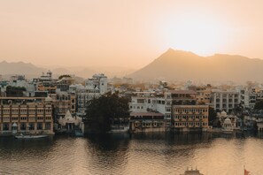 A riverside view of a city with buildings, a boat, and a mountain in the background. - Dreamyard Udaipur - Hostel (Udaipur)
