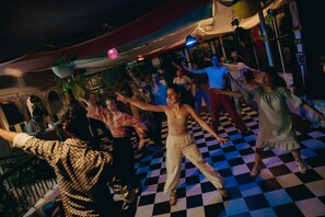 A group of people dancing on a checkered floor in a room with a red canopy and hanging plants. - Dreamyard Udaipur - Hostel (Udaipur)