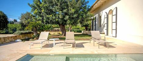 A pool area with clear water, surrounded by lounge chairs and a large tree.