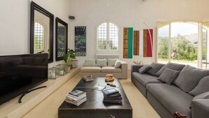 Un salon avec un téléviseur, des canapés, une table basse avec des livres et de grandes fenêtres offrant une vue dégagée.