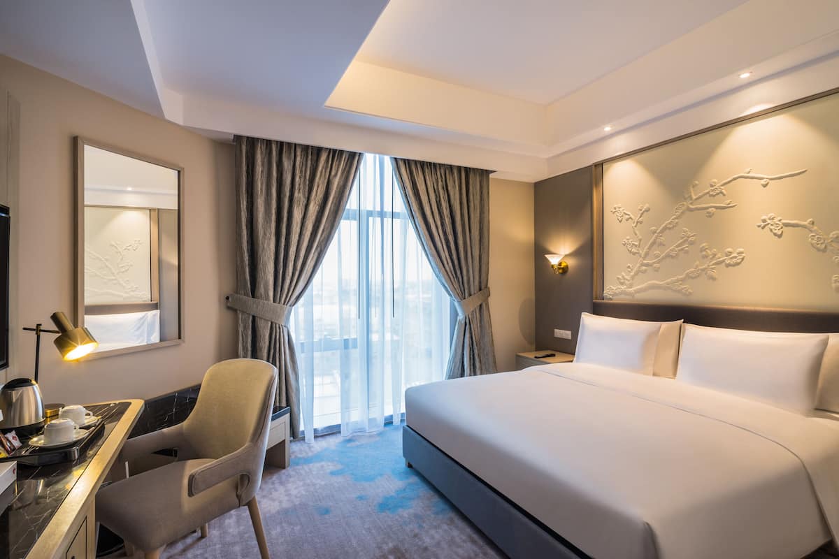 A hotel room with a large bed, a desk with a chair, a desk lamp, and a view of the outdoors through the window.