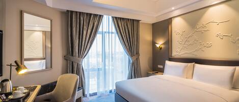 Habitación de hotel con una cama grande, un escritorio con silla, una lámpara de escritorio y vistas al exterior a través de la ventana.