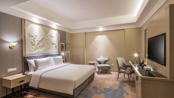 A modern hotel room with a large bed, a desk with a flat-screen TV, a seating area with two chairs, and a decorative wall panel.