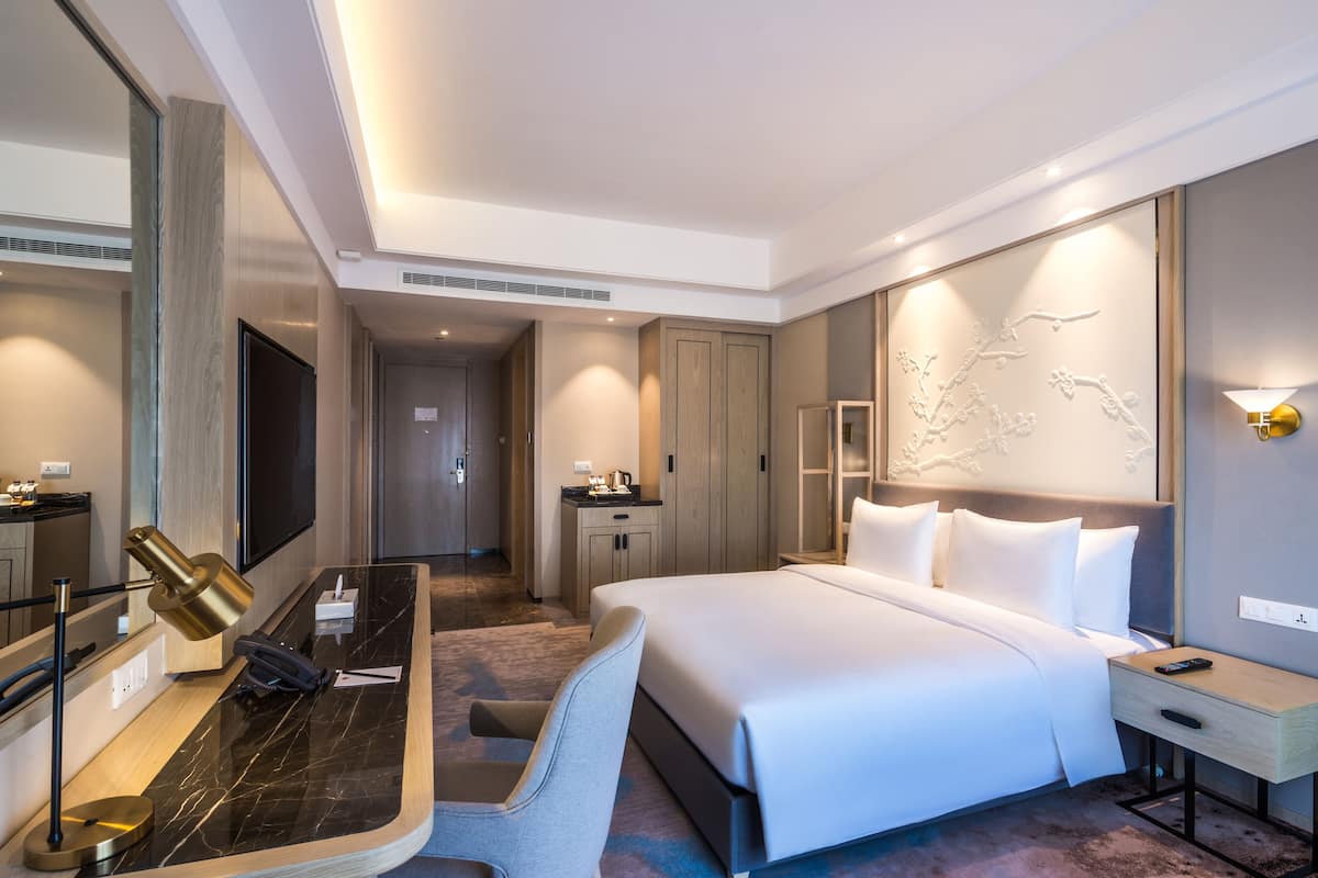 A modern hotel room with a large bed, a desk with a lamp, a mirror, and a wall-mounted artwork.