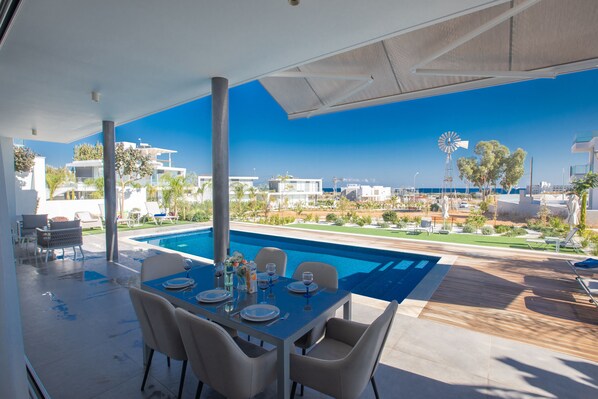 A modern outdoor dining area with a pool, overlooking a coastal landscape with buildings and a windmill.