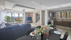 Uma sala de estar moderna com sofá, mesa de jantar para dois e área de cozinha visível por uma porta aberta.