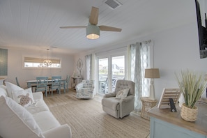 A living room with a white ceiling, a ceiling fan, a sofa with a turtle pillow, a chair, a dining table, and a view through the window. - "Unleashed & Untied" - Pet Friendly Canal Home, 3 Bedrooms / 2 Bathrooms (Holden Beach)