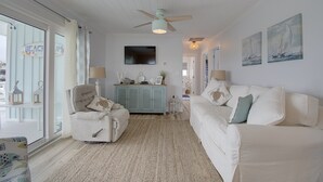 A spacious living room with a white sofa, a matching armchair, and a blue cabinet. A ceiling fan and a flat-screen TV are mounted on the wall.