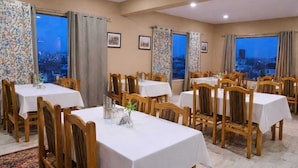 A dining area with multiple tables and chairs, large windows with curtains, and framed pictures on the walls. - Hotel Malik Residency (Leh)