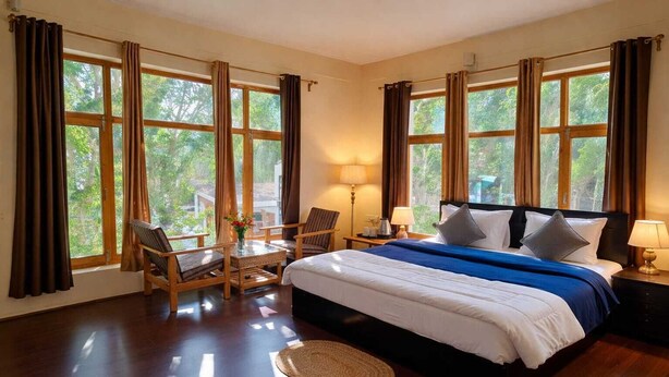 A spacious bedroom with a large bed, a sitting area with a table and chairs, and multiple windows offering a view of greenery. - Hotel Malik Residency (Leh)