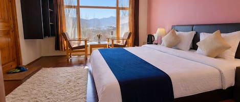 A hotel room with a large bed, a desk, two chairs, a TV, and a window with a view of mountains.