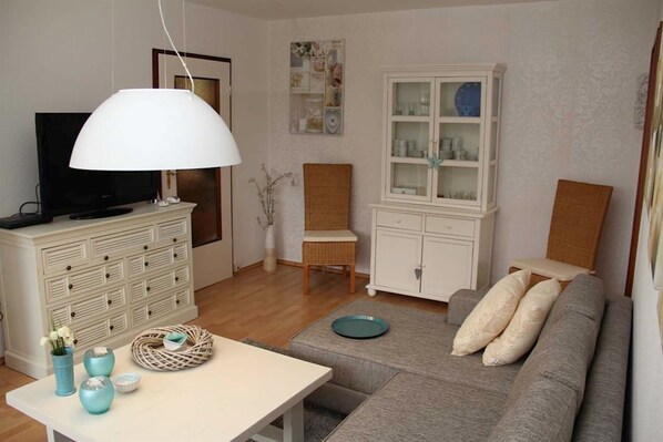 Un salon comprenant un canapé, un téléviseur, un meuble blanc, un fauteuil en osier et une table à manger agrémentée d’objets décoratifs.