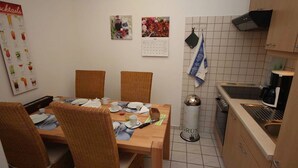 Ein Essbereich mit einem Holztisch, gedeckt für vier Personen, umgeben von Korbstühlen. An der Wand befinden sich ein Kalender und eine Cocktailkarte.