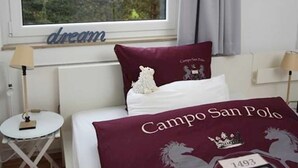 Ein Schlafzimmer mit großem Fenster, einem Bett mit burgunderfarbener Decke, einem kleinen Tisch mit Lampe und einem an der Wand befestigten Kalender.