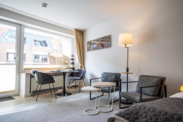 A hotel room with a bed, two chairs, a small table, a lamp, and a view of buildings outside.
