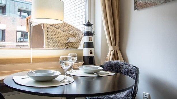 A dining area with a round table, two white plates, two wine glasses, a lamp, a lighthouse model, and a view of a brick building through a window.