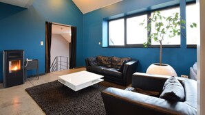 Una sala de estar moderna con chimenea, sofá de cuero negro, mesa de centro blanca y un ventanal con vistas a la vegetación.