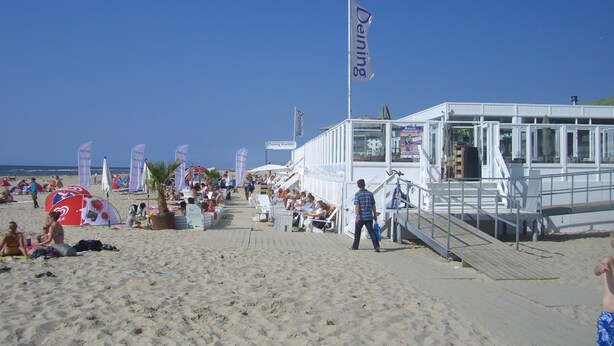 A beachside building with a glass facade, a deck area, and multiple flags. - Charming Home by the Lake (West-Grafdijk)