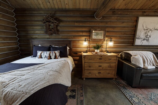 A log cabin bedroom with a bed, a dresser, a chair, and a wall-mounted light fixture.