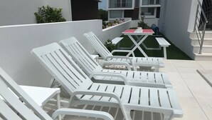 En rad vita solstolar är uppställda på en kakelbelagd uteplats, med ett litet bord och ett blått föremål i närheten.