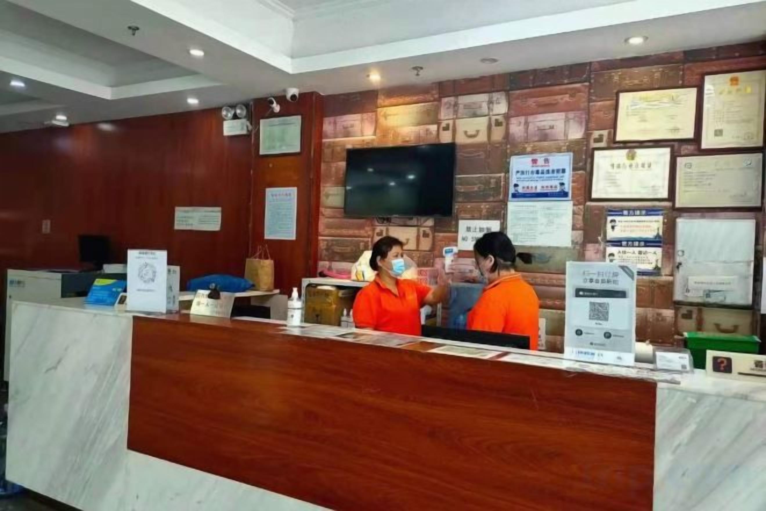 A hotel reception desk with a wooden finish, a flat-screen TV mounted on the wall, and various certificates and notices displayed.