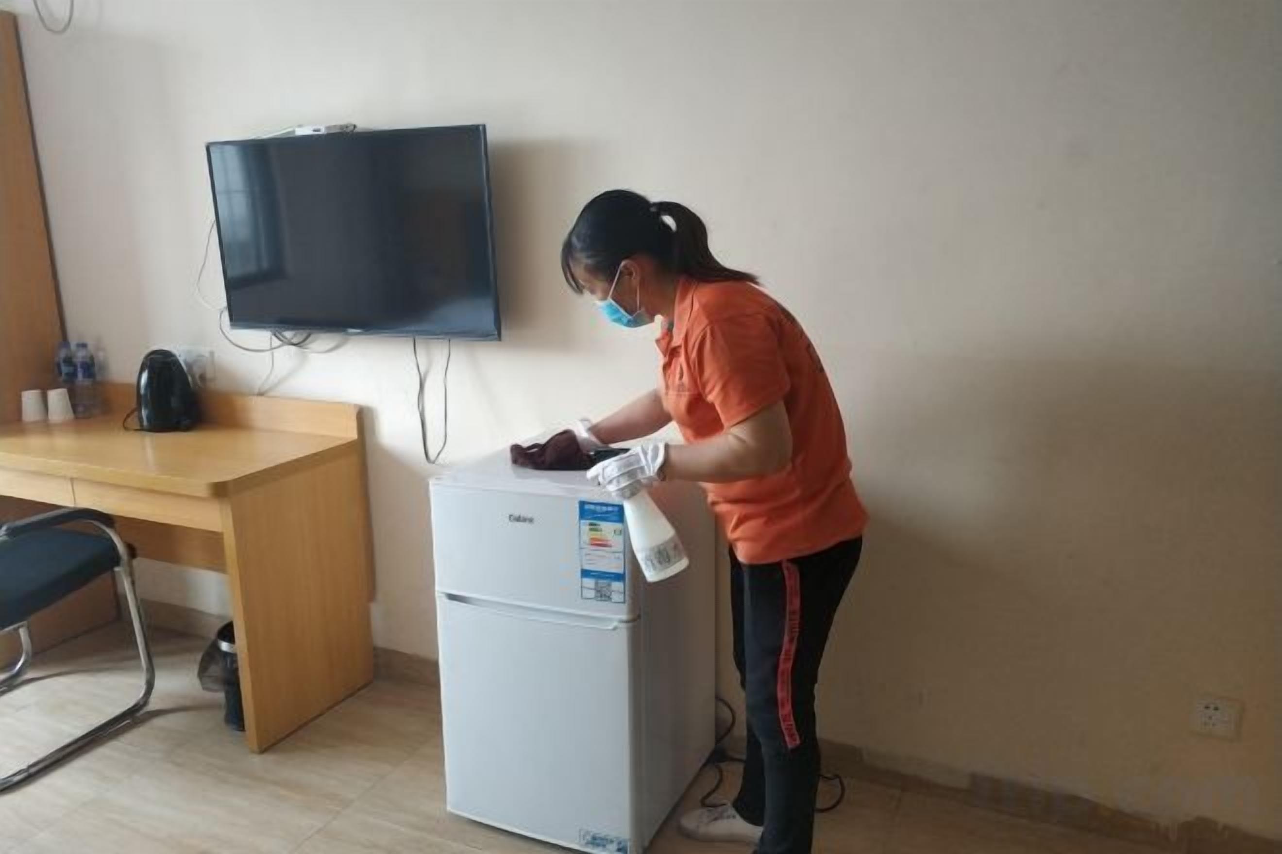 A person cleaning a white refrigerator in a room with a wooden desk, a chair, and a television mounted on the wall.
