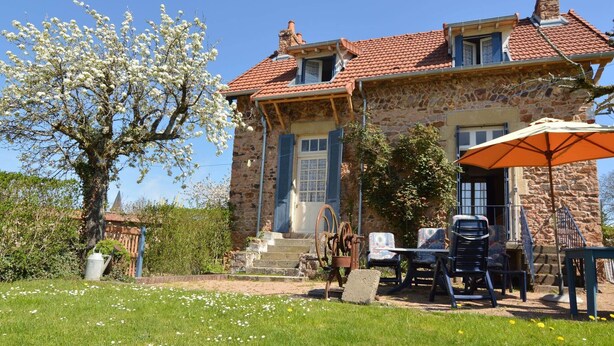 Ein Steinhaus mit rotem Ziegeldach, eine Holztreppe zum Eingang, Sitzgelegenheiten im Freien und ein Baum mit weißen Blüten.