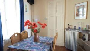 Ein Essbereich mit einem Tisch für zwei Personen, einer Vase mit roten Blumen, Korbstühlen, einer Heizung und einem Gemälde an der Wand.