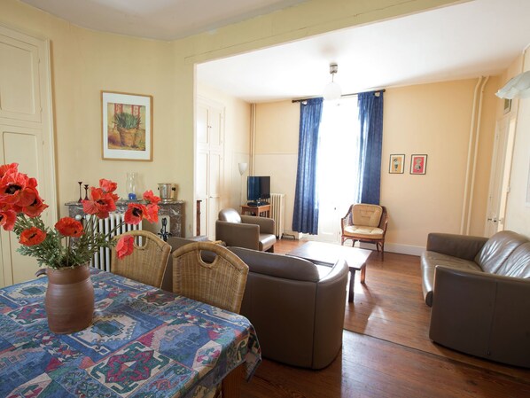 Un salon comprenant un canapé, des fauteuils, une table basse et une table à manger recouverte d’une nappe à motifs. On y trouve une cheminée ornée d’un vase rempli de fleurs rouges, un téléviseur et un tableau encadré accroché au mur.