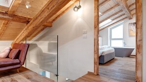 A modern loft bedroom with exposed wooden beams, a glass partition, a bed, and a large window.