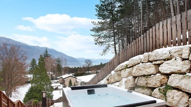 A hot tub with a stone wall and wooden fence, surrounded by a snowy landscape and trees.