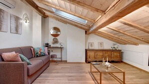 A loft-style room with a sloped ceiling, wooden beams, a sofa with cushions, a round mirror, a wooden cabinet, and a glass coffee table.