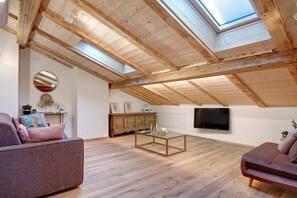 Habitación en ático con vigas de madera, tragaluz, sofá, mesa de centro, televisor y armario de madera.