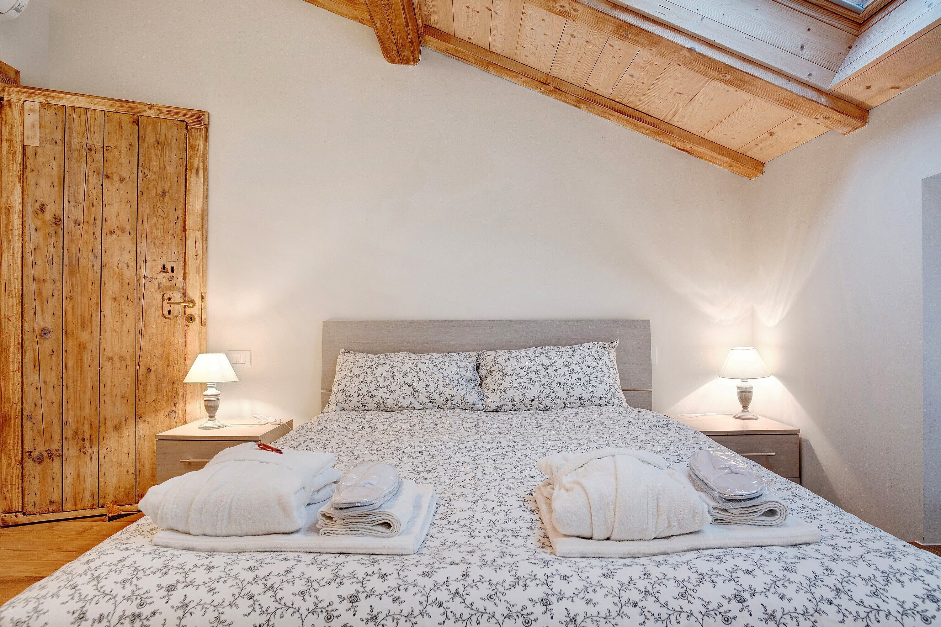 Une chambre avec une armoire en bois, un lit avec une literie à motifs floraux, deux tables de chevet avec des lampes et une lucarne.