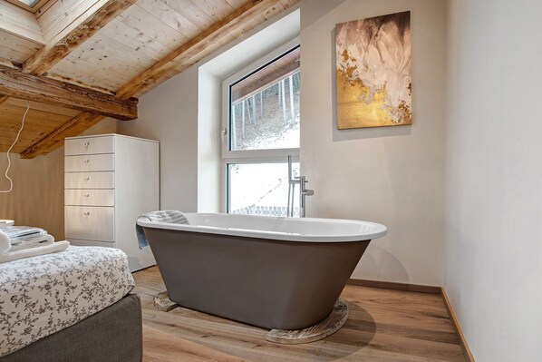 A modern bathroom with a freestanding bathtub, a wooden ceiling, and a large window.