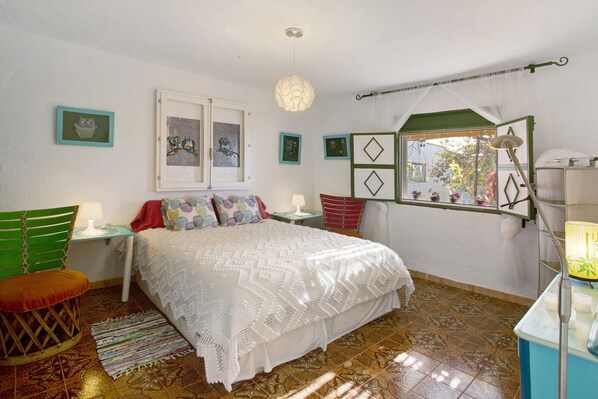 A bedroom with a bed, a window with a view, a chair, a table, and a lamp.