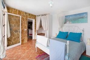 A bedroom with a stone wall, a bed with blue pillows, a white headboard, and a painting of a pier.