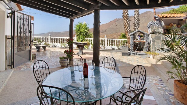 Une table avec un plateau en verre, des chaises, une bouteille et des verres, le tout sur une terrasse offrant une vue sur les montagnes.