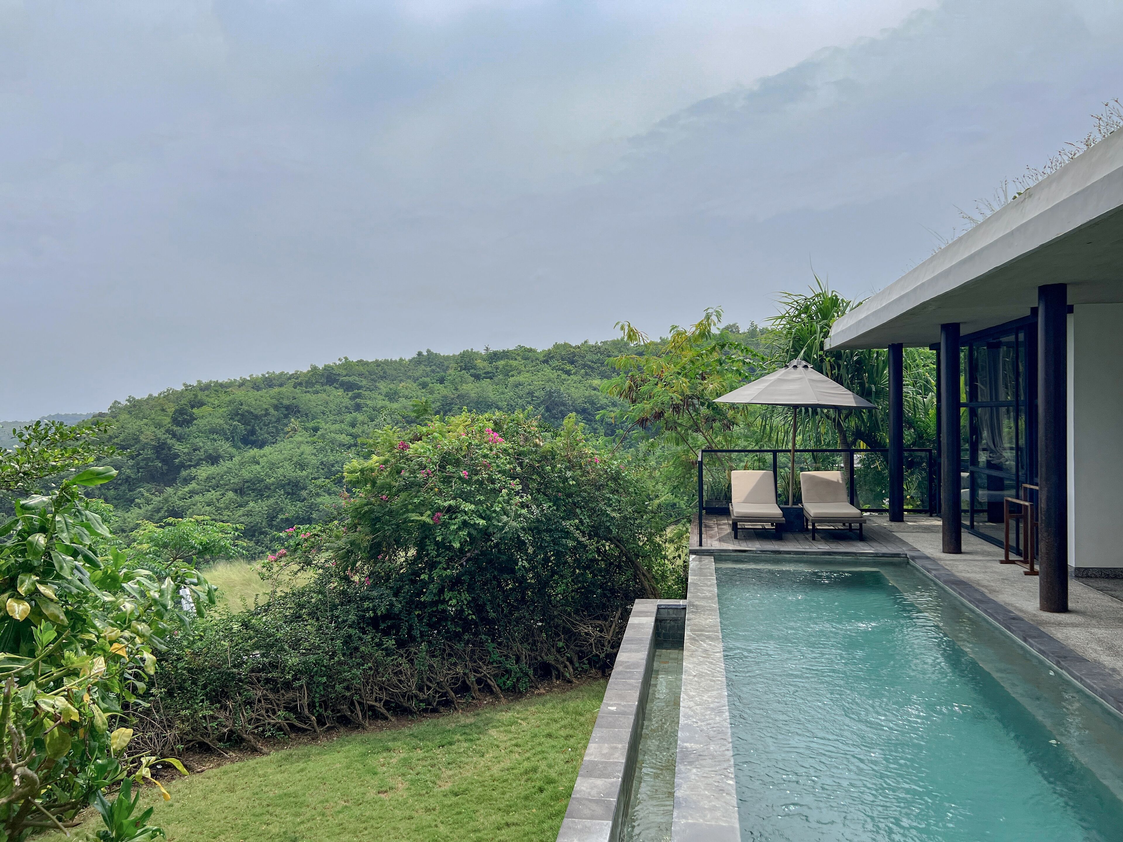 A pool with a view of a lush green hillside, a covered seating area with chairs, and a modern building with large windows.